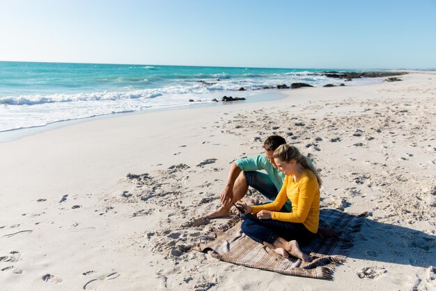 Vista lateral de una pareja caucásica reclinada en la playa con el cielo azul y el mar en el fondo, abrazando y mirando su teléfono inteligente