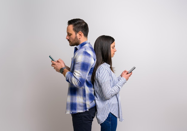 Vista lateral de una pareja joven seria de pie espalda con espalda y mensajes de texto a través de teléfonos inteligentes