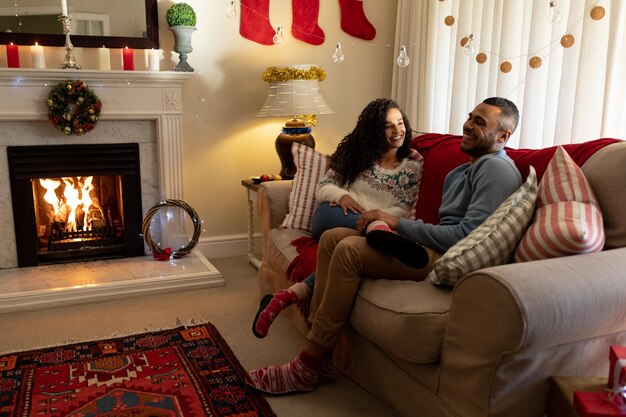 Foto vista lateral de una pareja de raza mixta en su sala de estar en navidad, sentada en un sofá hablando y sonriendo