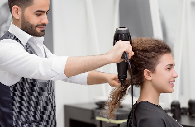 Vista lateral del peinado árabe que seca el cabello rizado de una clienta sonriente.