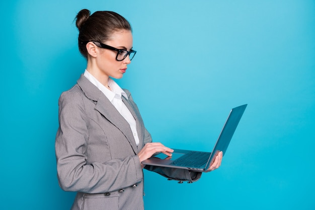 Vista lateral de perfil de atractiva dama ocupada enfocada sosteniendo en la mano usando un portátil aislado sobre fondo de color azul brillante