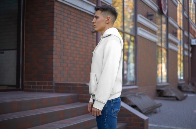 Vista lateral de una presentación de sudadera con capucha blanca sobre un chico joven en el contexto de un edificio de ladrillo. Maqueta de ropa casual para publicidad en tienda. Plantilla de ropa para su patrón y diseño.