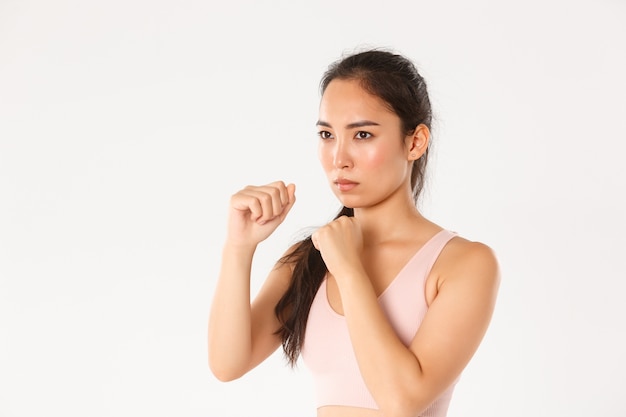 Vista lateral de primer plano de una chica asiática seria en ropa de fitness levantando los puños cerrados y frunciendo el ceño determinado, yendo a clases de defensa personal, boxeo de sombras.