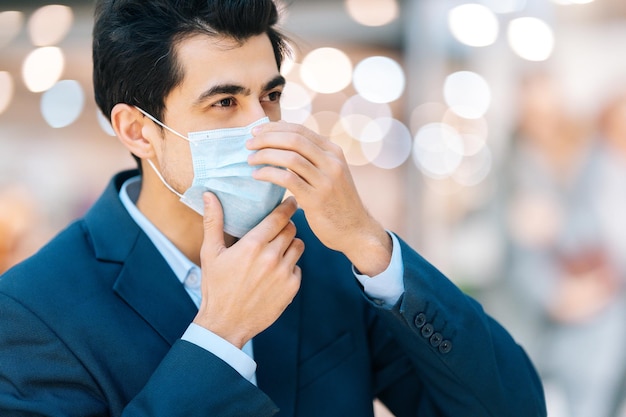 Vista lateral de primer plano de un joven confiado en un traje elegante con mascarilla de protección mirando hacia otro lado en el pasillo del centro comercial fondo borroso Primer plano de un hombre guapo aplicando máscara