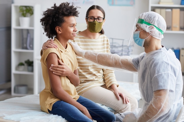 Foto vista lateral retrato de un adolescente afroamericano sonriendo después de la vacunación contra la covid en la clínica con un enfermero felicitándolo