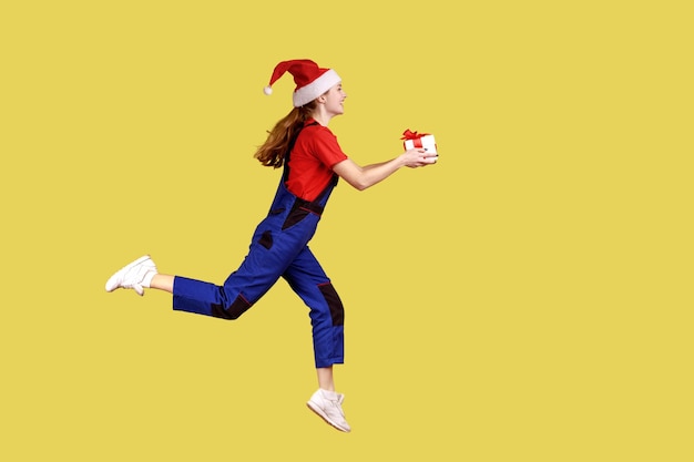 Vista lateral retrato completo da mulher de correio correndo com caixa de presente para entregar o Natal para os clientes, vestindo macacão azul e chapéu de papai noel. Tiro de estúdio interior isolado em fundo amarelo.