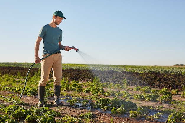 Vista lateral retrato de cuerpo entero de un joven trabajador regando los cultivos en la plantación de hortalizas y sonriendo mientras está de pie al aire libre contra el cielo azul, espacio de copia