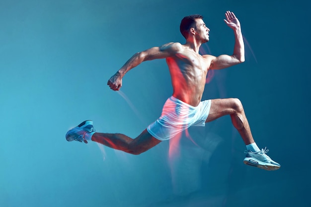 Vista lateral del retrato de cuerpo entero del tipo de fitness corriendo a un joven medio desnudo con pantalones cortos blancos en azul