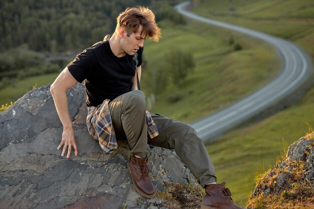 Vista lateral retrato de guapo chico caucásico sentado en la cima de la montaña relajante después de escalar, vestido informalmente, en la roca. Camino En El Fondo. Viaje, viaje, concepto de aventura