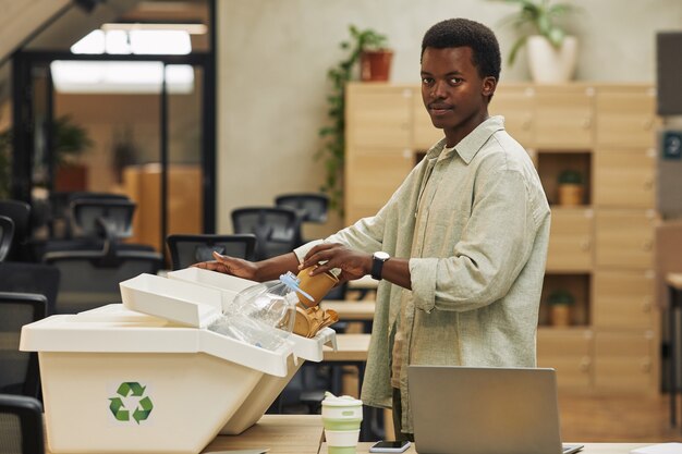 Vista lateral retrato de joven afroamericano poniendo vaso de papel en la papelera de clasificación de residuos en la oficina, espacio de copia