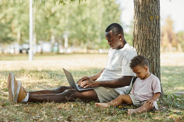 Vista lateral retrato de joven afroamericano usando laptop al aire libre en el parque con lindo bebé hijo policía ...