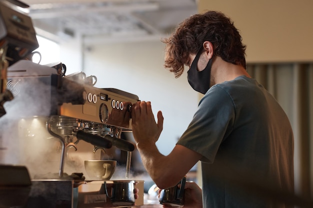 Foto vista lateral retrato de joven barista con máscara mientras prepara café recién hecho en la cafetería o cafetería, espacio de copia