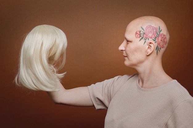 Vista lateral retrato de mujer calva confiada con peluca de cabello rubio sobre fondo marrón en estudio, conciencia de alopecia y cáncer, espacio de copia