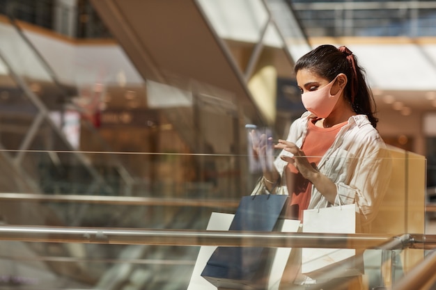 Vista lateral retrato de mujer joven con máscara mientras hace compras en el centro comercial y usa el teléfono inteligente, espacio de copia