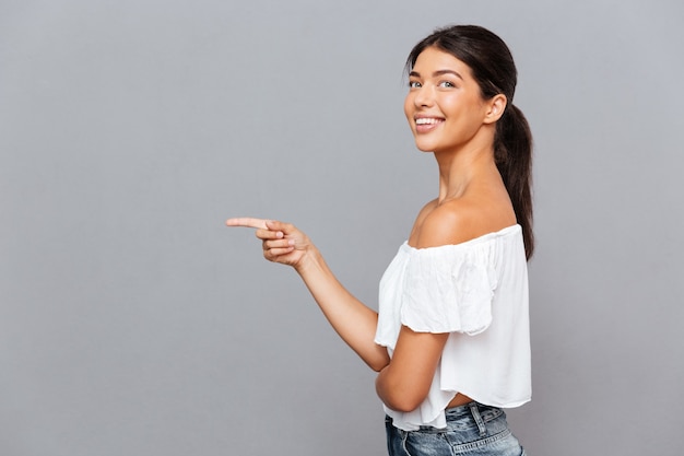 Vista lateral retrato de una mujer joven sonriente que señala el dedo a un lado aislado en la pared gris