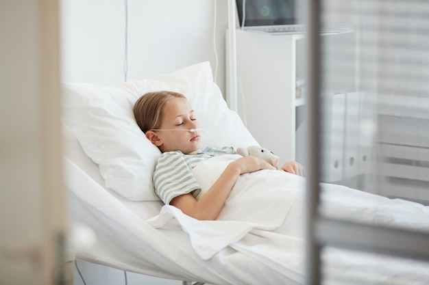 Vista lateral retrato de niña tendida en la cama de un hospital con soporte de oxígeno, espacio de copia