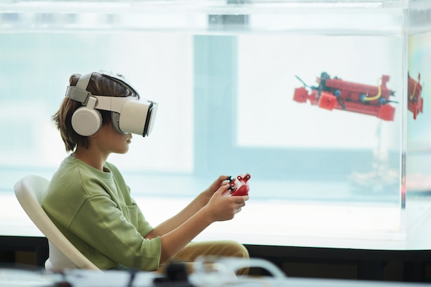 Vista lateral retrato de niño usando auriculares VR mientras opera un barco robótico en el laboratorio de la escuela, espacio de copia