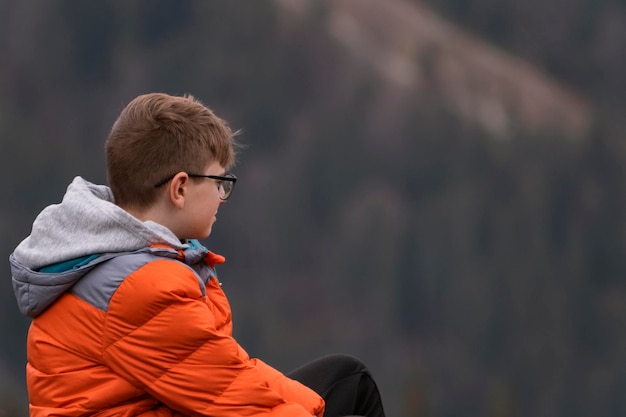 Vista lateral retrato de niño viste chaqueta deportiva naranja y gafas Chico con cabello castaño al aire libre Vista de las montañas