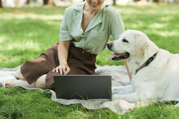 Vista lateral retrato de perro blanco tumbado sobre la hierba verde en el parque con una mujer joven con ordenador portátil en el fondo, espacio de copia