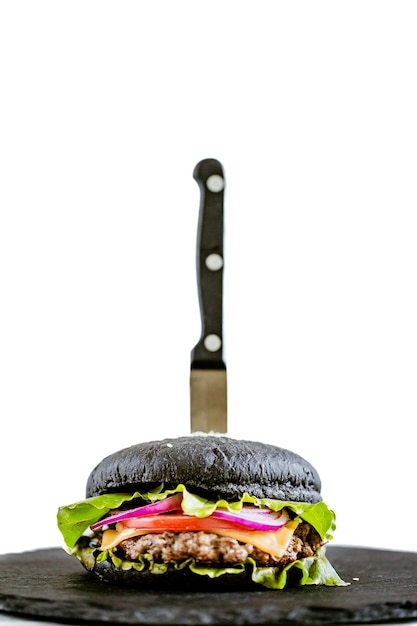 Foto vista lateral de la sabrosa hamburguesa de ternera negra con verduras en tablero de piedra con cuchillo sobre fondo blanco.