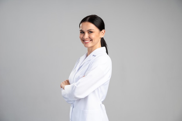Foto vista lateral de la sonriente doctora en bata de laboratorio con los brazos cruzados mirando a otro lado y posando sobre fondo gris. concepto de medicina