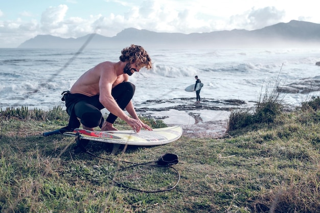 Vista lateral de un surfista masculino sonriente, descalzo y con el torso desnudo, en cuclillas y poniendo cera en una tabla de surf en la costa del océano ondulado