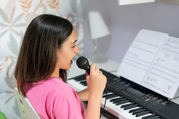 Vista lateral de la talentosa jovencita con micrófono interpretando una canción y tocando el piano eléctrico durante el tiempo libre en casa