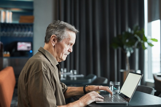 Vista lateral del trabajador autónomo senior ocupado serio con bigote sentado en la mesa negra en el café y realizando tareas en la computadora portátil