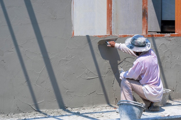 Foto vista lateral del trabajador constructor asiático enyesando la pared de cemento en el sitio de construcción de la casa