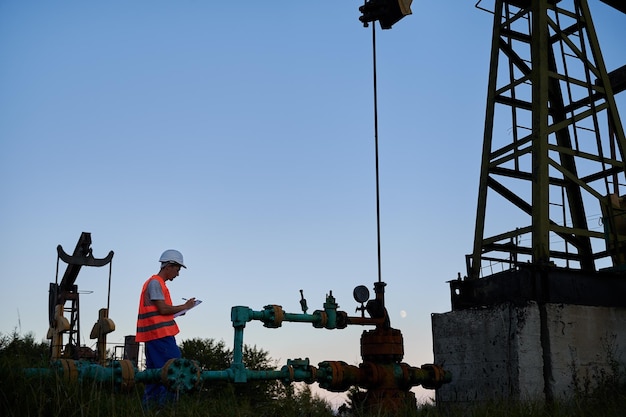 Vista lateral de un trabajador de pozo de petróleo que escribe los resultados de la inspección de la tubería en el clipboard Ingeniero tomando notas mientras realiza el servicio de las actividades de la plataforma petrolera contra el cielo azul