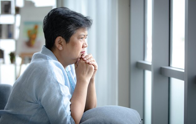 Vista lateral de la triste mujer asiática madura con el pelo corto uniendo las manos y mirando por la ventana en casa durante el día