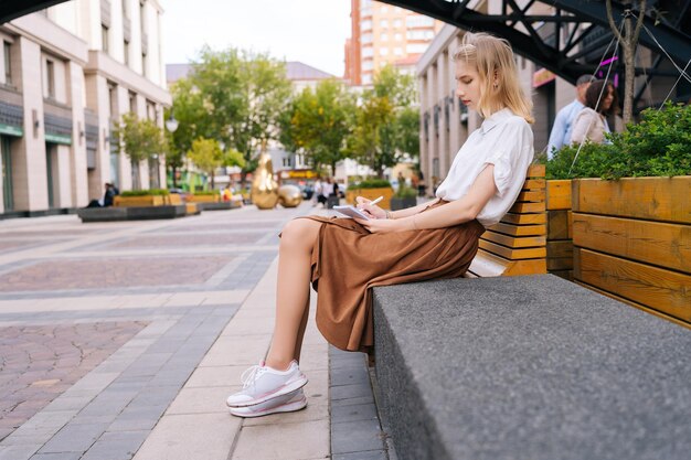 Vista lateral de una turista pensativa haciendo notas de aventura de viaje sentada en un banco en la plaza del centro Mujer muy hábil dibujando en un cuaderno de bocetos