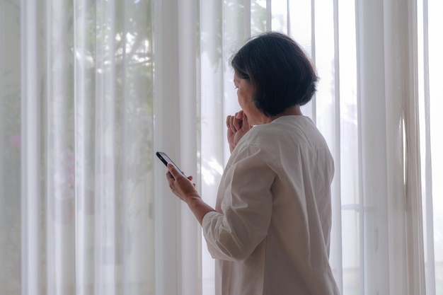 Vista lateral Uma mulher segurando um telefone celular em pé perto da janela roendo as unhas