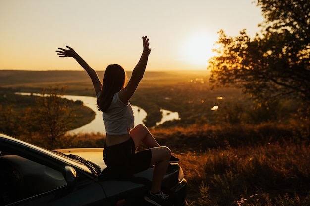 Vista lateral del viajero joven con las manos en alto mirando la hermosa puesta de sol mientras está sentado en el coche.