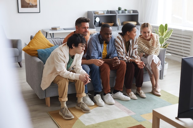 Vista de longitud completa en un grupo multiétnico de amigos viendo la televisión en casa juntos mientras están sentados en una fila en el sofá, copie el espacio