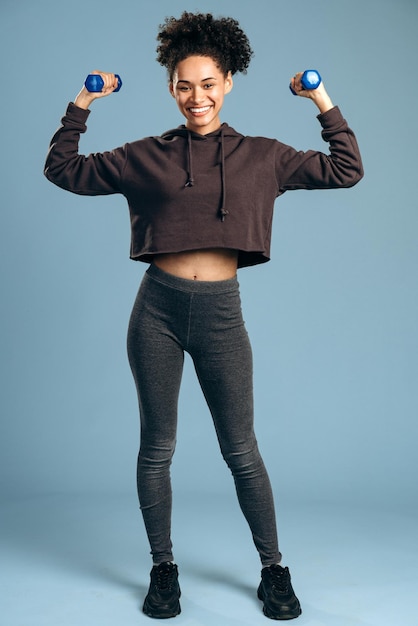 Vista de longitud completa de la hermosa chica deportiva multirracial sosteniendo pesas y sonriendo mientras posa aislado sobre fondo azul. Foto de stock