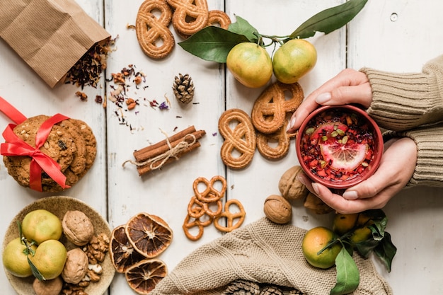 Vista de manos femeninas sosteniendo la taza con bebida caliente entre galletas caseras, nueces, clementinas y especias en el cuadro blanco