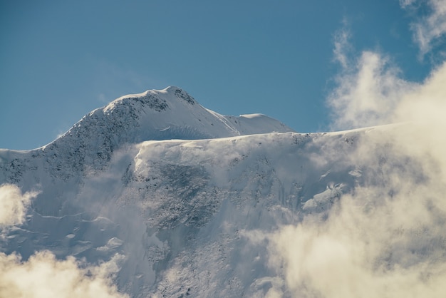 Vista minimalista de la pared de la montaña cubierta de nieve en espesas nubes bajas bajo el sol. Pintoresco paisaje de montaña brillante con pico de nieve blanca entre densas nubes en el cielo azul. Maravilloso paisaje con cima nevada.