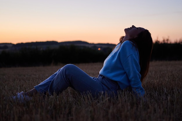 Una vista de una mujer sentada con una sonrisa en la pradera y mirando hacia arriba durante la puesta de sol