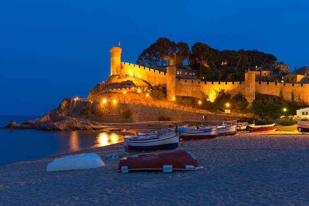 Vista nocturna de la fortaleza y los barcos de pesca y la niña sentada en la playa de gran platja y la playa de badia de tossa...