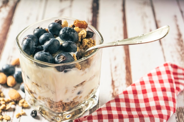 Vista normal de um copo com iogurte, mirtilo e parfait de granola em uma mesa de madeira rústica. Conceito de alimentação saudável e natural.