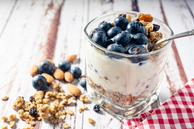 Vista normal de um copo com iogurte, mirtilo e parfait de granola em uma mesa de madeira rústica. Conceito de alimentação saudável e natural.