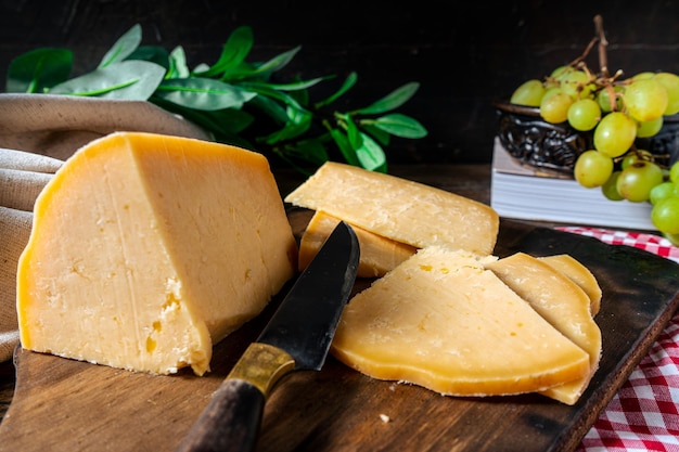 Vista normal de un trozo de queso provolone cortado sobre una tabla y unas uvas al fondo.