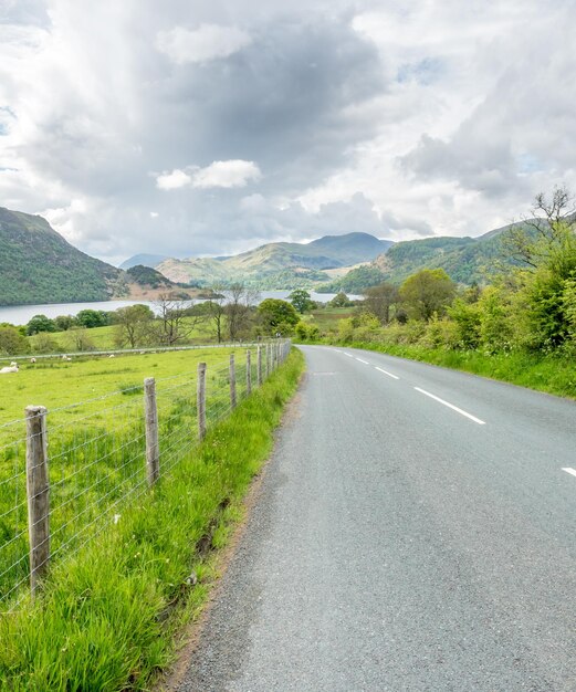 Vista del paisaje de campo de árboles verdes y hierba con montaña a lo largo de un camino estrecho con cerca de alambre de púas bajo el cielo nublado de la noche en la Inglaterra rural