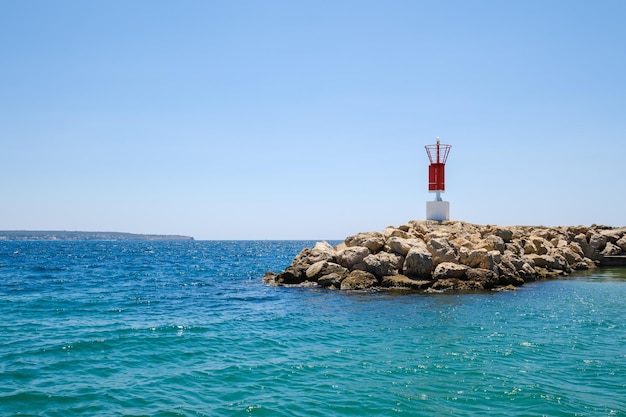 Vista panorámica de la baliza roja situada en la costa rocosa bañada por agua de mar turquesa bajo un cielo azul sin nubes