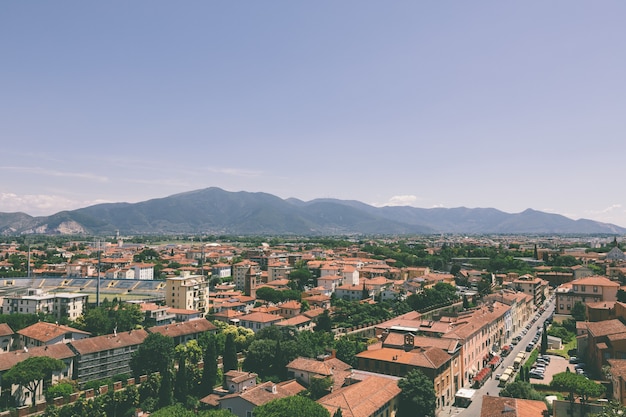 Vista panorámica de la ciudad de Pisa con edificios históricos y montañas lejanas desde la Torre de Pisa. Día de verano y cielo azul soleado.