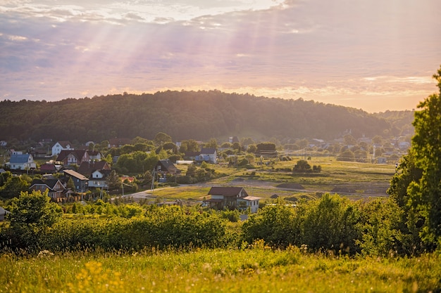 Vista panorâmica da montanha até um pequeno povoado rural entre campos e florestas ao pôr do sol no verão.