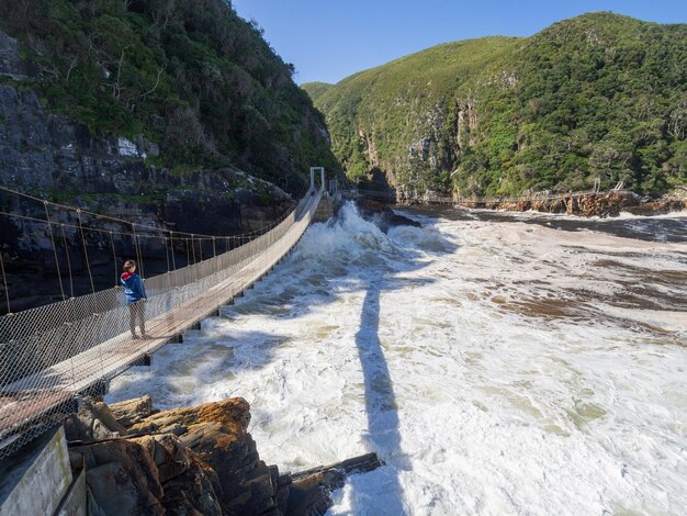 Vista panorâmica de uma jovem caminhando sobre uma ponte de pé suspensa sobre o oceano na foz do rio storms, na áfrica do sul