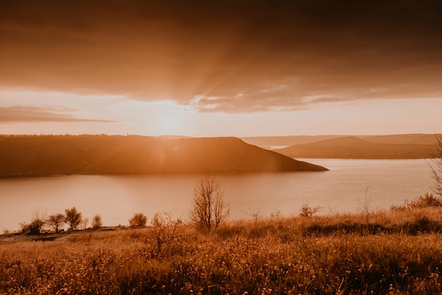 Vista panorâmica desde a falésia até ao grande rio, lago, mar à distância, silhuetas das ilhas, pôr do sol laranja ardente