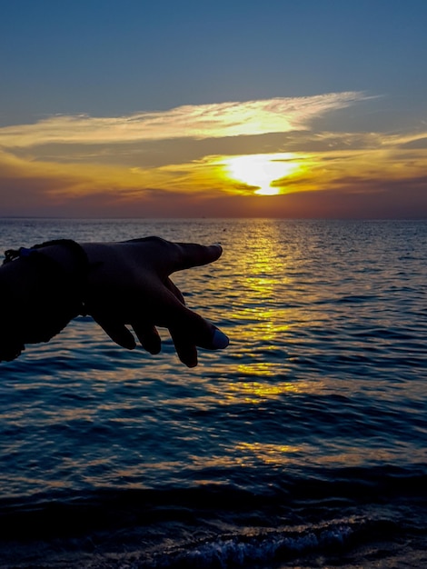 Foto vista panorâmica do mar contra o céu durante o pôr-do-sol com a mão apontando para o pór-do- sol